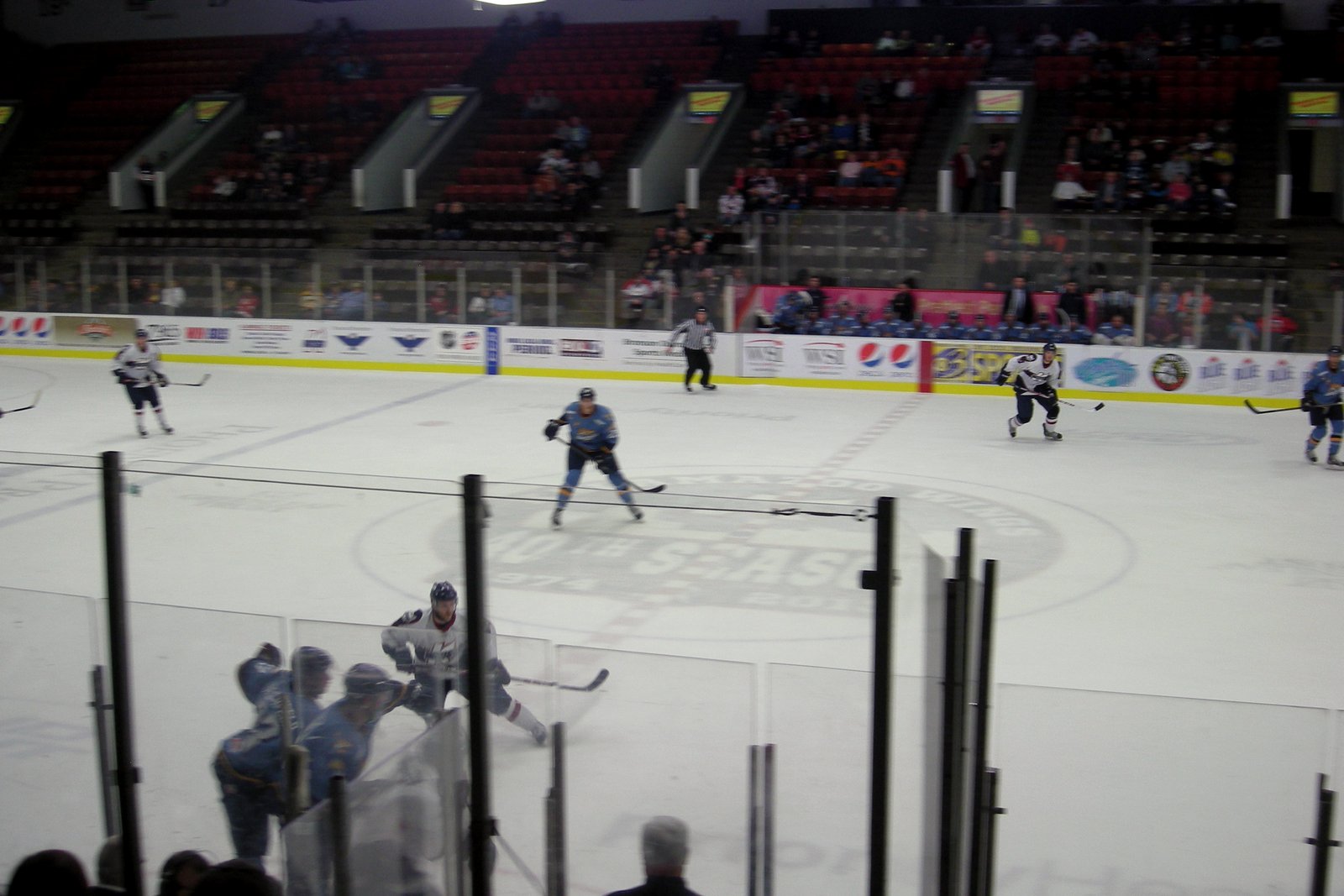 Hockey players in white and in light blue jerseys skate near center ice during a game in an arena with red seats, seen through the glass behind the players’ bench.