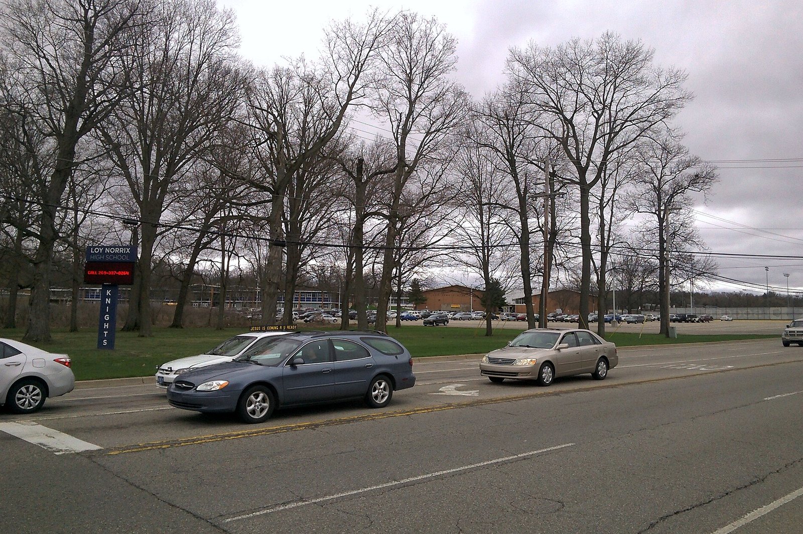 A wide suburban road with cars passing in front of a grassy lawn and tall bare trees, a blue Loy Norrix Knights sign at left and a low brick school building in the background under a cloudy sky.