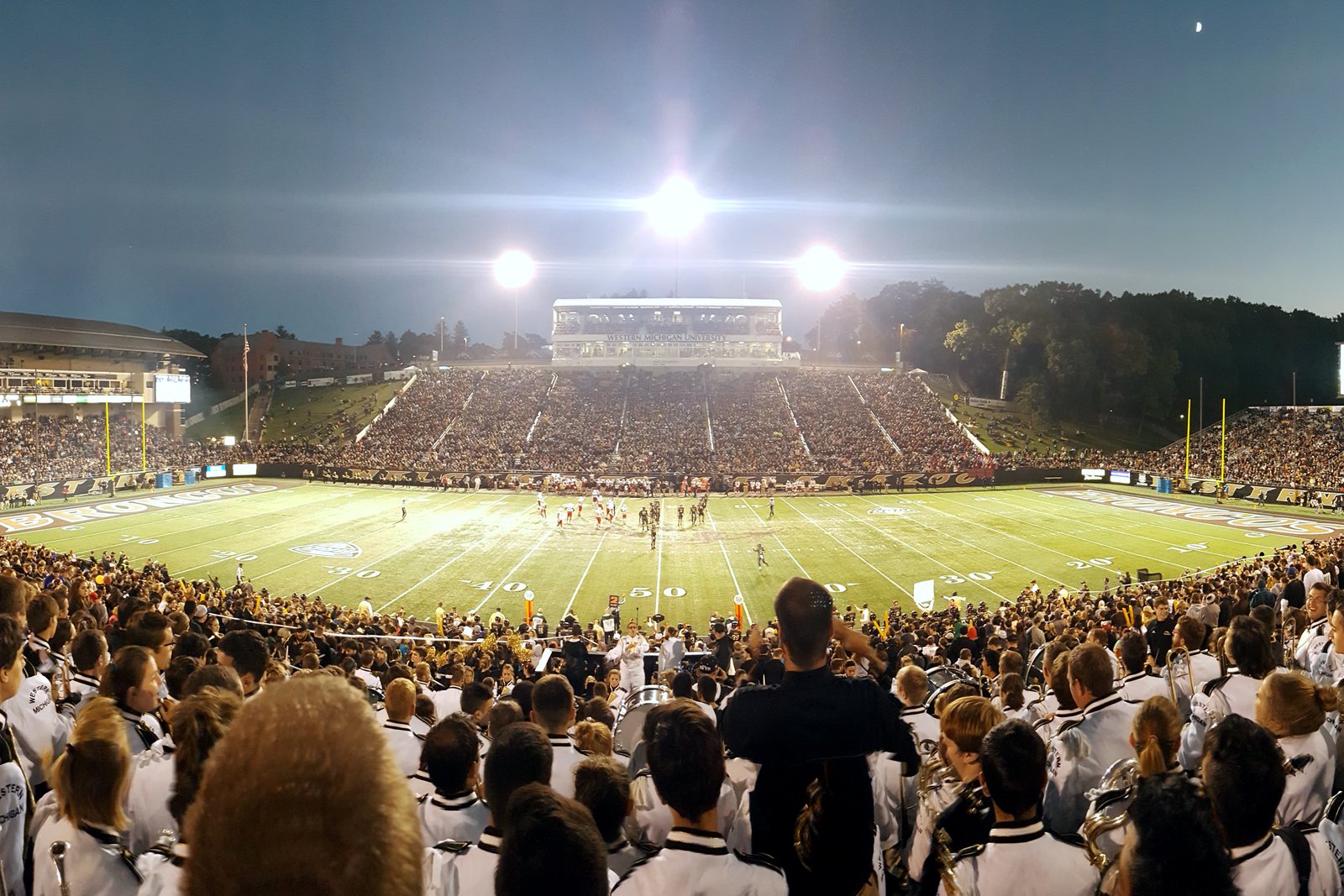 A panorama at dusk of a packed college football stadium from the top of the student section, with band members in white uniforms in the foreground and lights over the field.