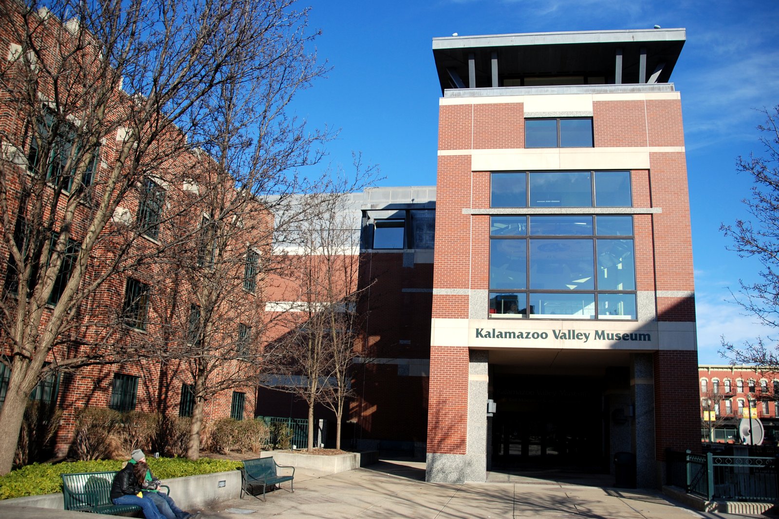 A red brick building with a tall glass-fronted tower labeled Kalamazoo Valley Museum stands beside bare trees and a plaza with benches under a blue sky.