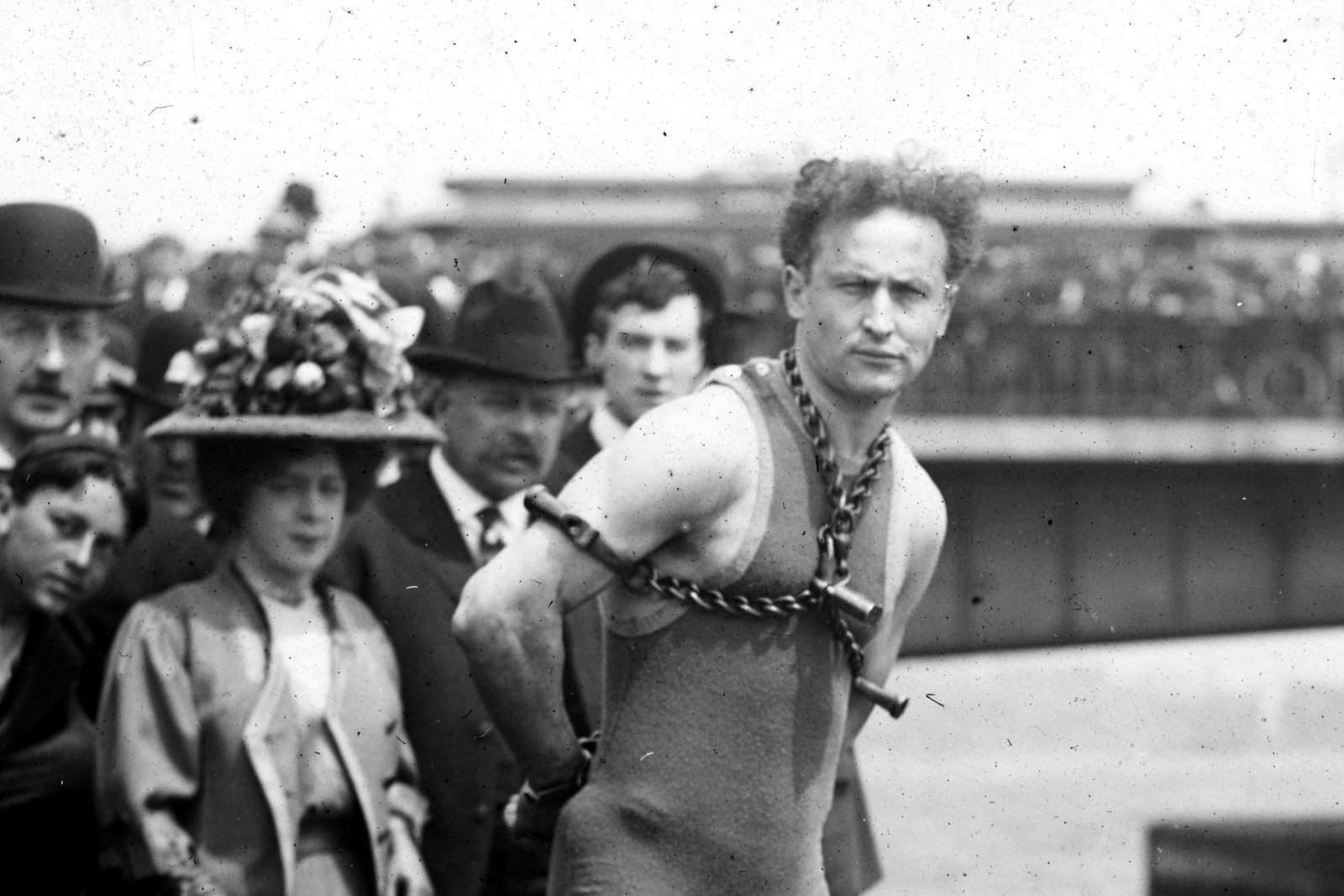 A black-and-white photo of a shirtless man in chains and handcuffs standing among a crowd of people in hats.