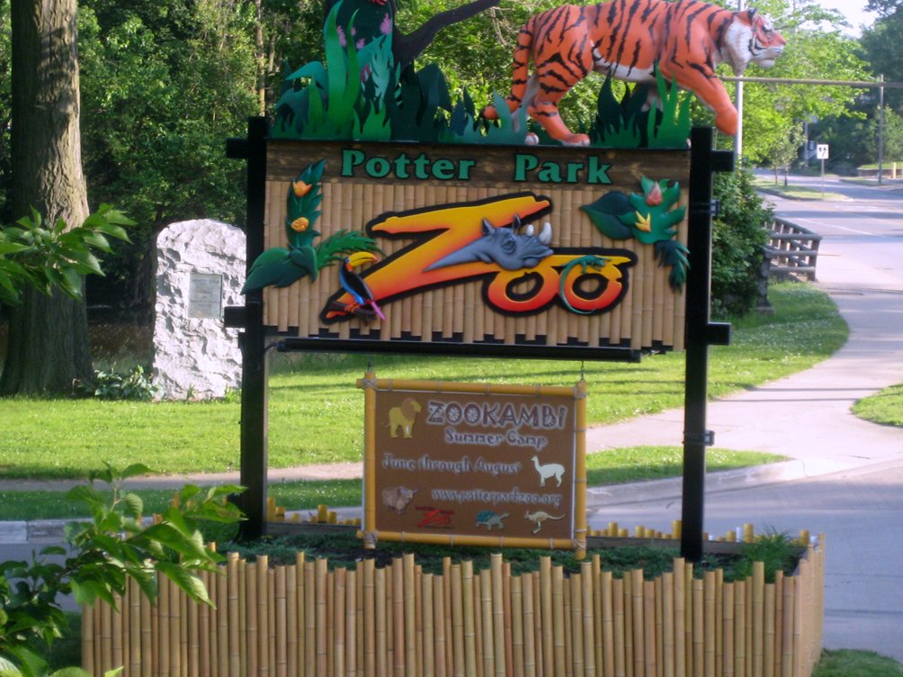 A colorful Potter Park Zoo entrance sign topped by a tiger sculpture and framed with bamboo, beside a curving road and green lawn under leafy trees.
