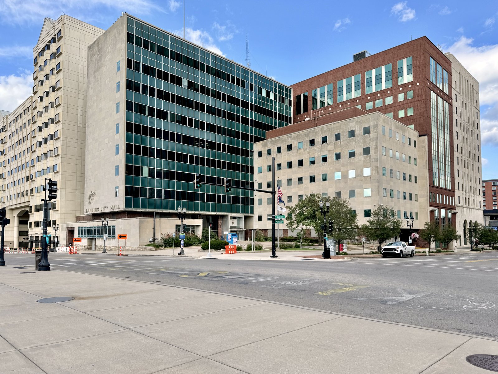 A ten-story modernist office tower with green glass panels and a lower limestone wing stand at a downtown Lansing intersection, with a manhole cover in the sidewalk-lined street in front.