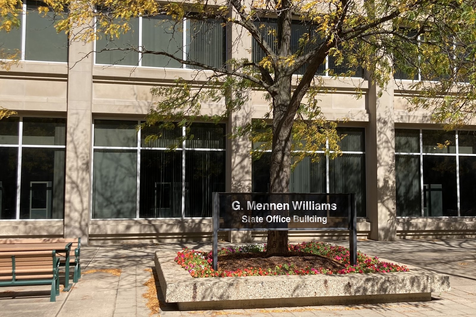 The entrance to the G. Mennen Williams State Office Building in Lansing, with a black sign bearing the building's name in a flower bed under a tree in front of tan stone and glass walls.