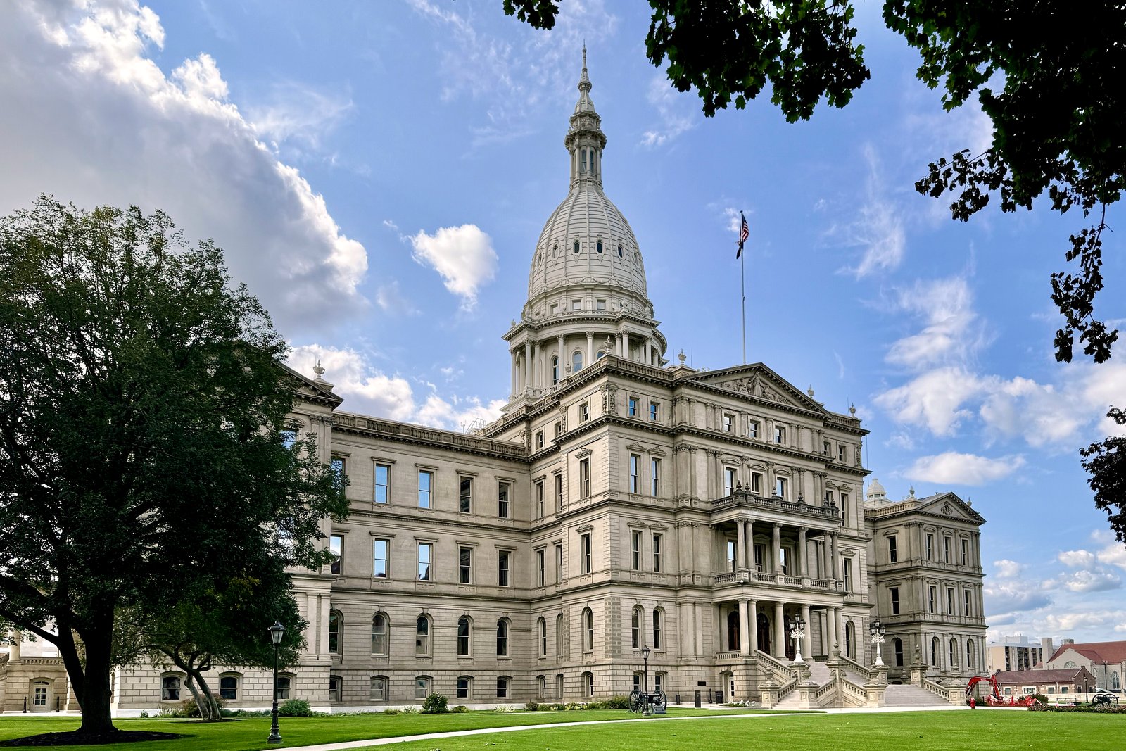 The Michigan State Capitol, a tan stone building with a tall white dome, behind a green lawn and large trees under a partly cloudy blue sky.