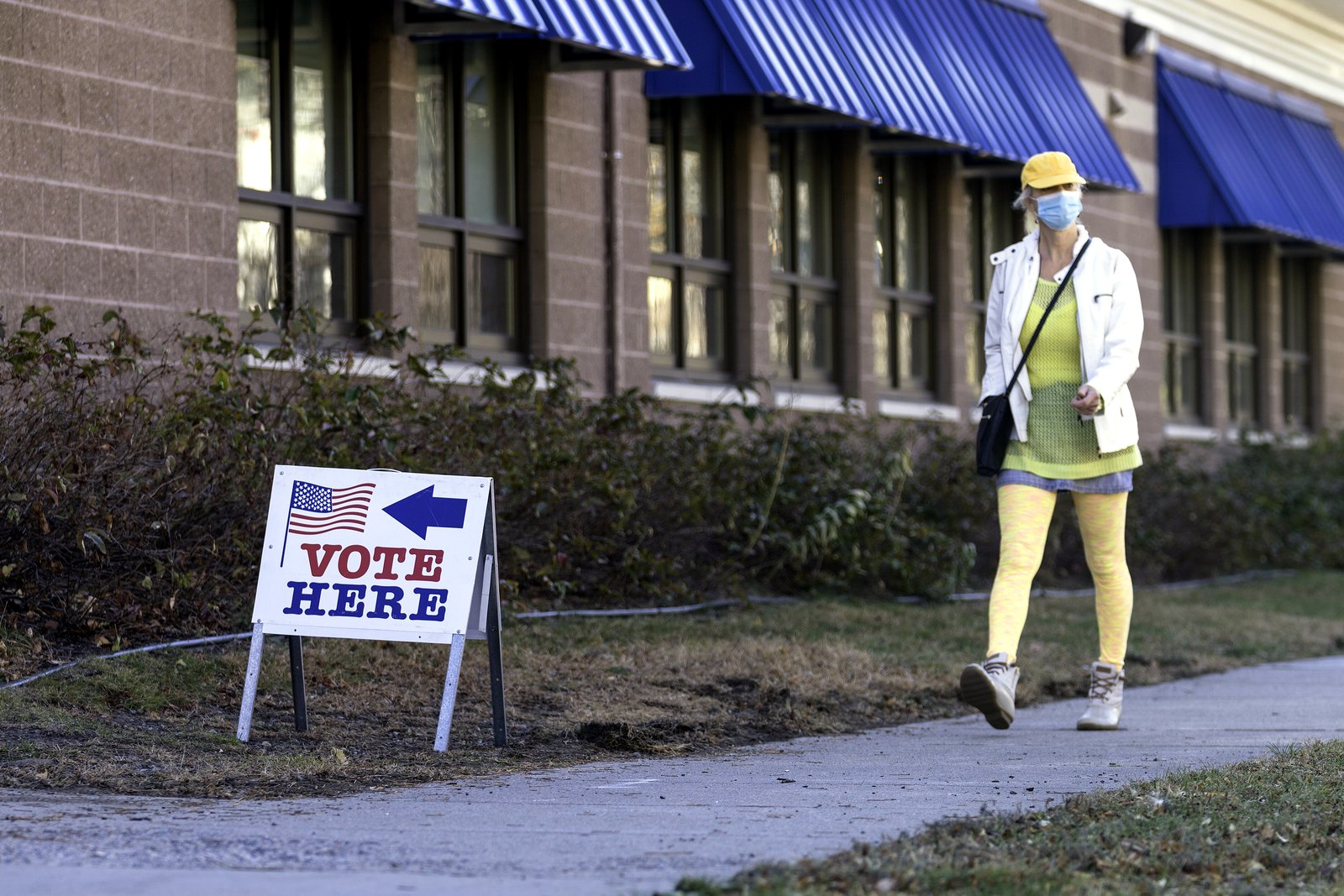 A Vote Here sign with an arrow on a lawn beside a brick building with blue awnings, as a woman in a mask walks past.