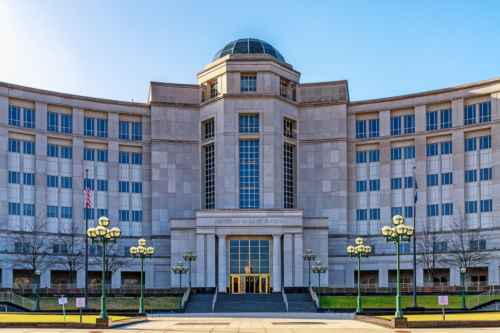 A large pale stone government building with a central rotunda and rows of windows behind a wide plaza with lampposts.
