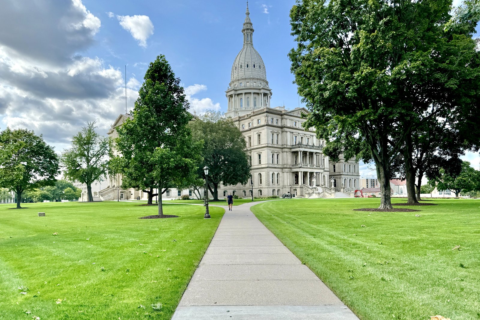 A domed white stone capitol building at the end of a long sidewalk lined with green lawns and tall trees on a sunny day.