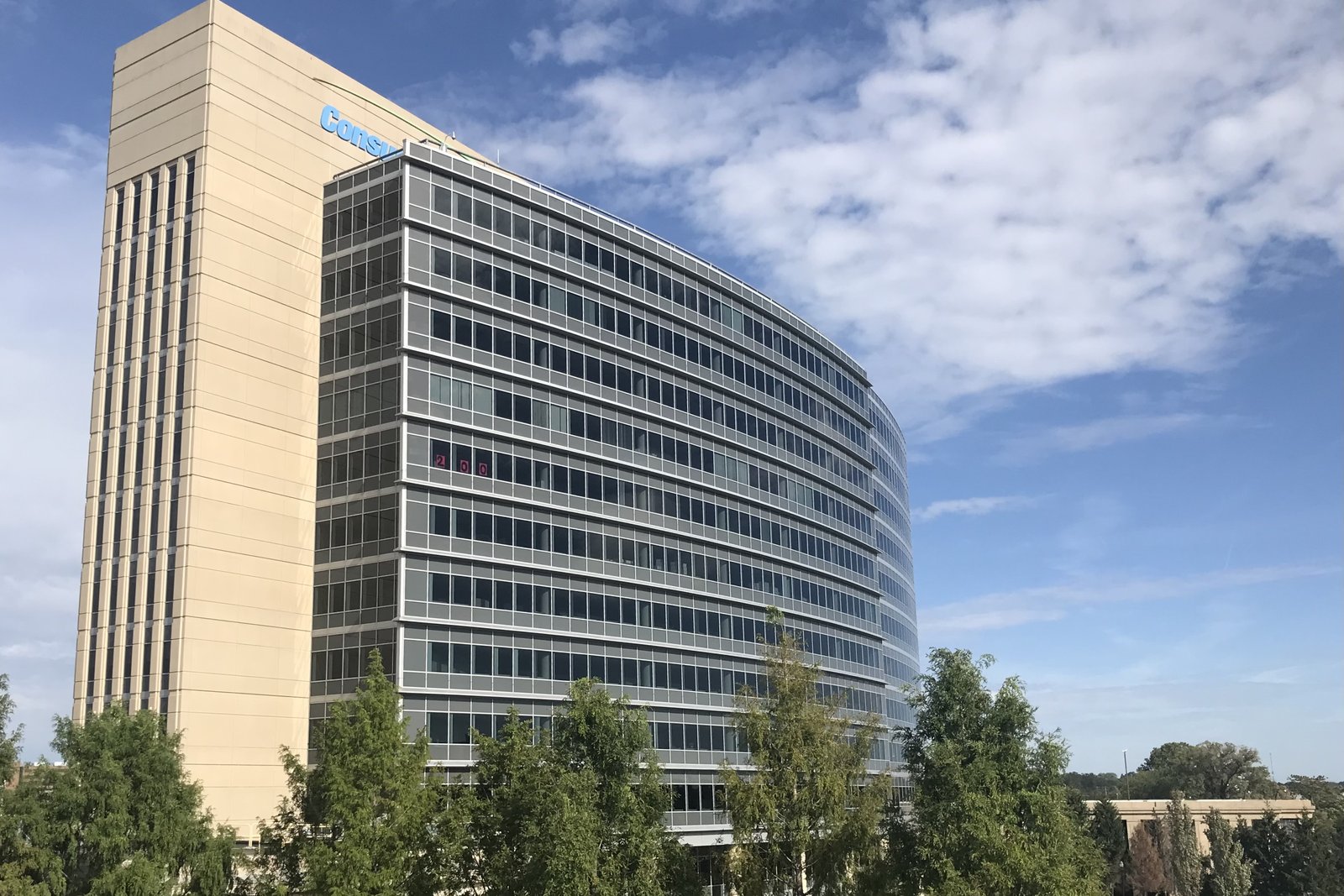 A tall modern office tower with horizontal bands of windows beside a lower brick building under a partly cloudy sky.