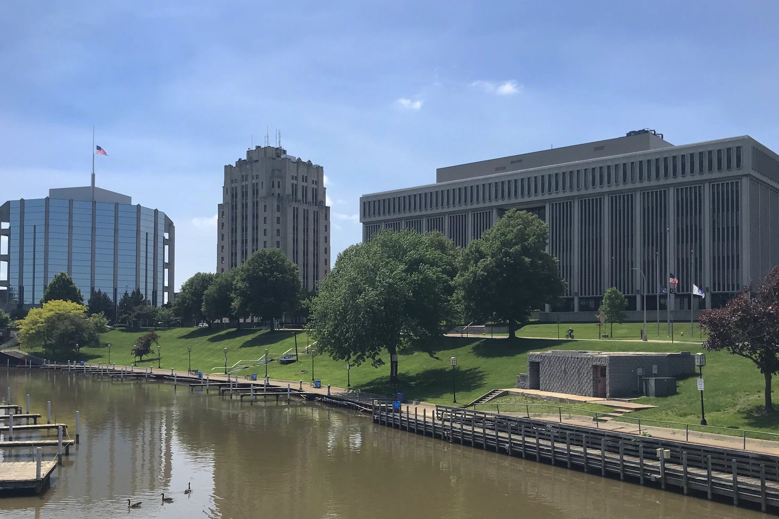 A riverfront in downtown Mount Clemens with boat docks and a grassy slope in front of a round blue-glass building flying an American flag, a 1930s stone tower and a long modern office building, under a blue sky.