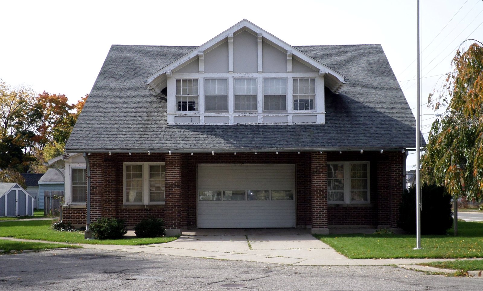 A brick, two-story bungalow-style firehouse with a white garage door and a flagpole sits on a corner lot under a gray sky.