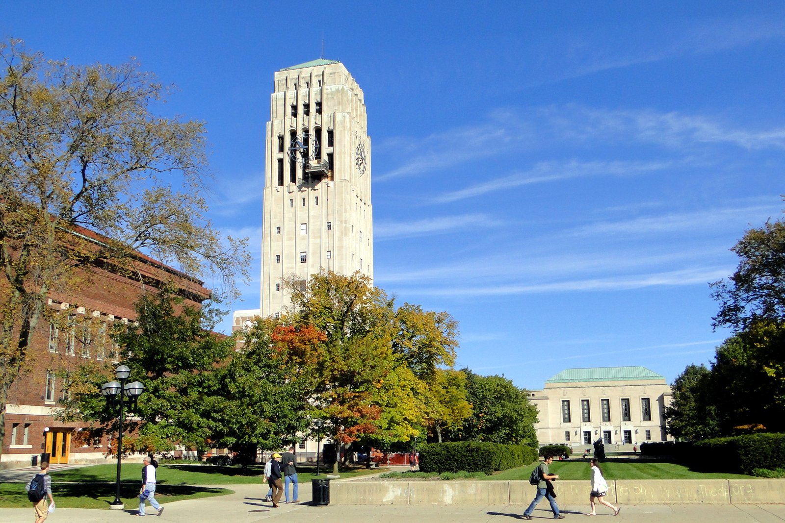 A tall, pale stone clock and bell tower rising above autumn trees with orange and yellow leaves, with people walking on a sidewalk under a blue sky.