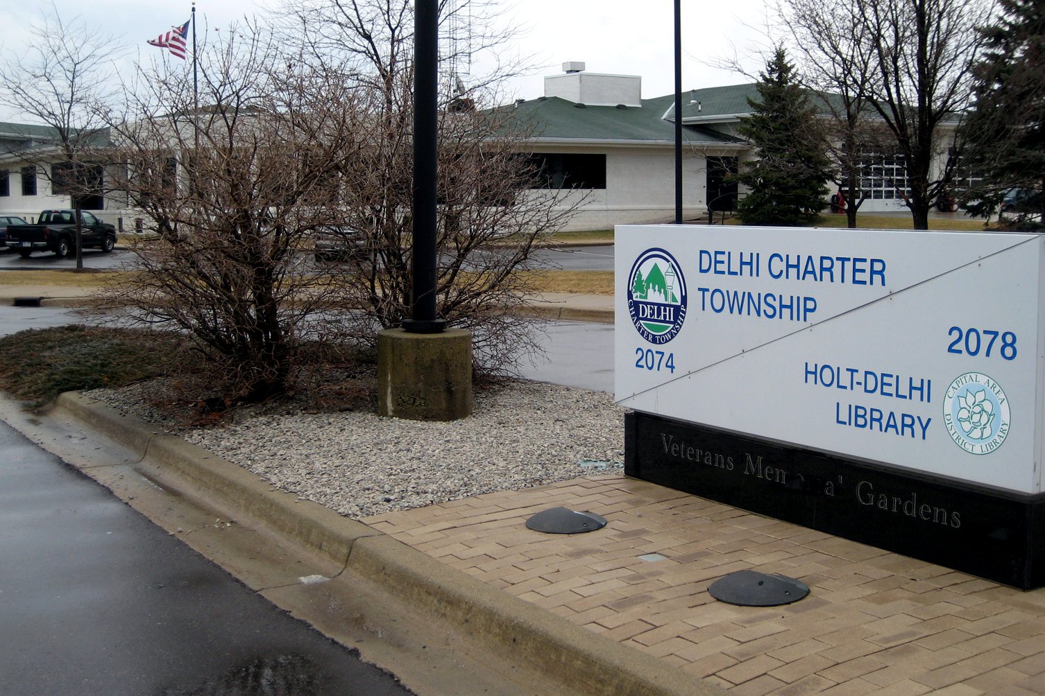 A Delhi Charter Township sign for the Holt-Delhi Library and administrative offices on a landscaped corner, with bare trees behind it.