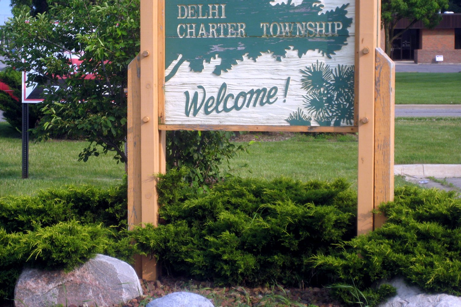 A wooden sign with green lettering reading Delhi Charter Township Welcome, set in a landscaped bed with shrubs, rocks and trees.