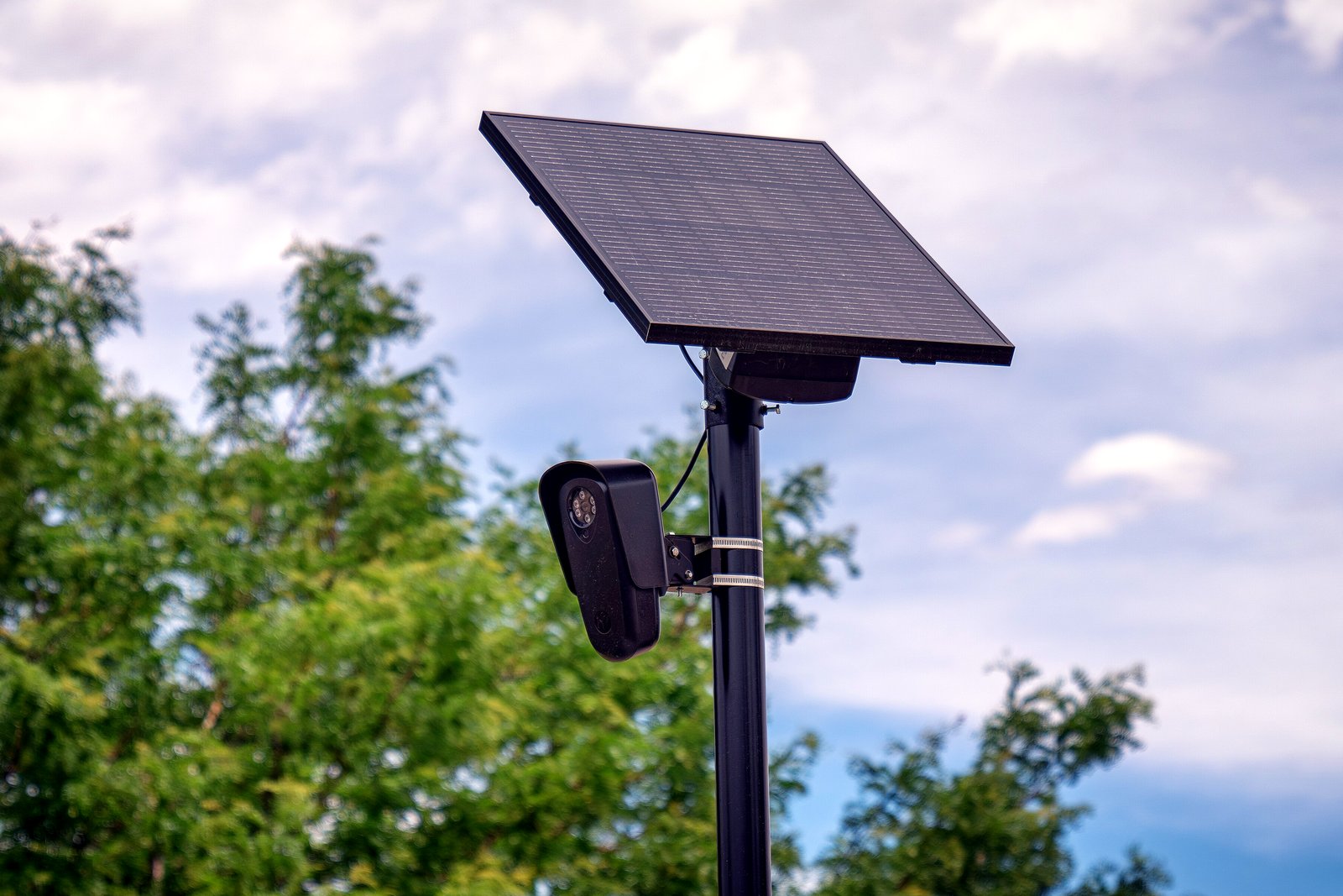 A black license plate reader camera and a solar panel mounted on a metal pole against a blue sky with clouds and trees.