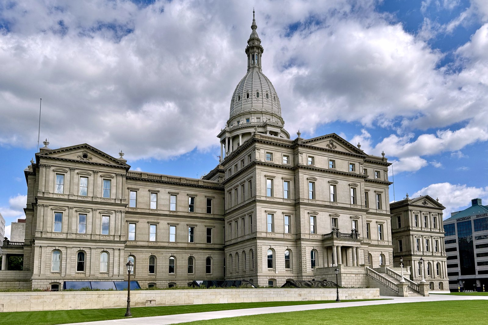 The Michigan State Capitol, a cream-colored Renaissance Revival building with a tall white dome, under a partly cloudy sky behind a green lawn.