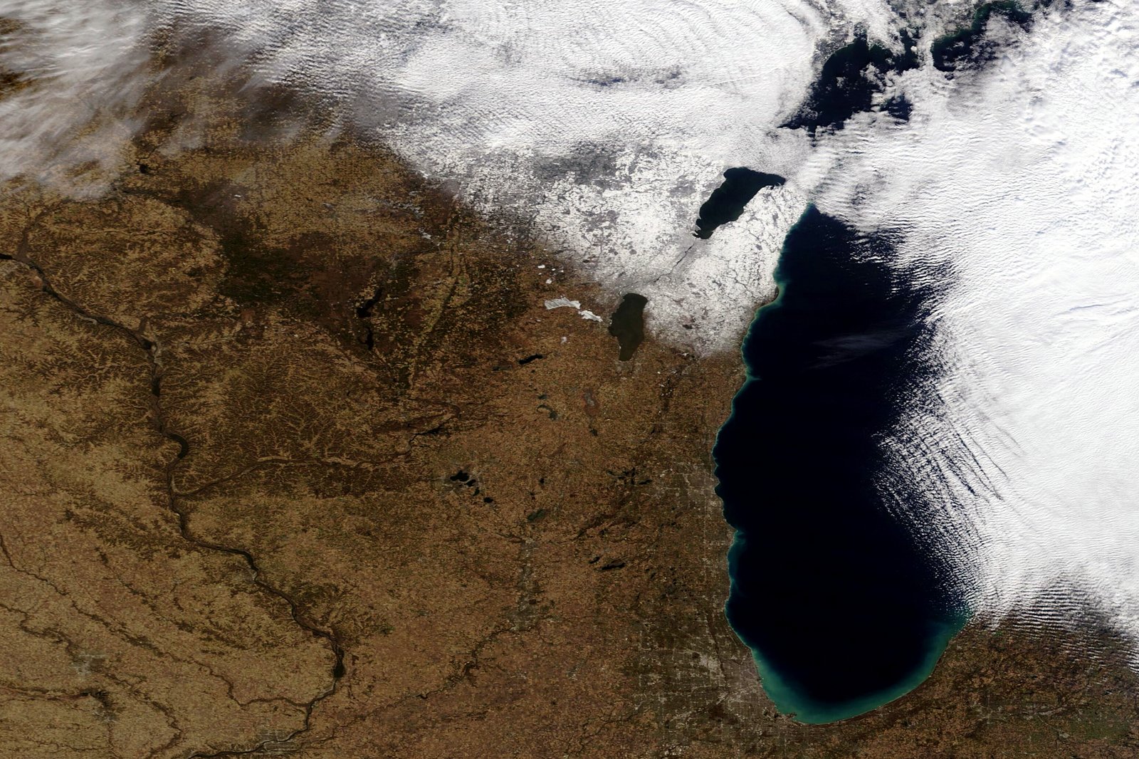 A satellite view of Lake Michigan, dark blue, with white bands of lake-effect clouds and snow streaming over Michigan and a brown, snow-free landscape to the west.