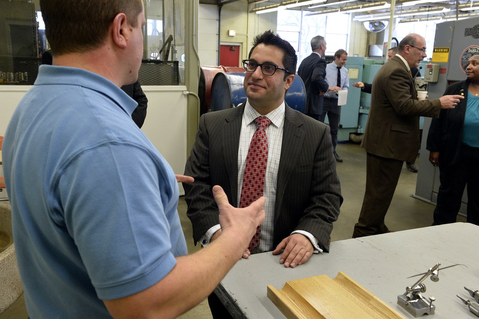A state lawmaker in a suit talks with a welding instructor in a prison vocational shop, with other visitors standing nearby.
