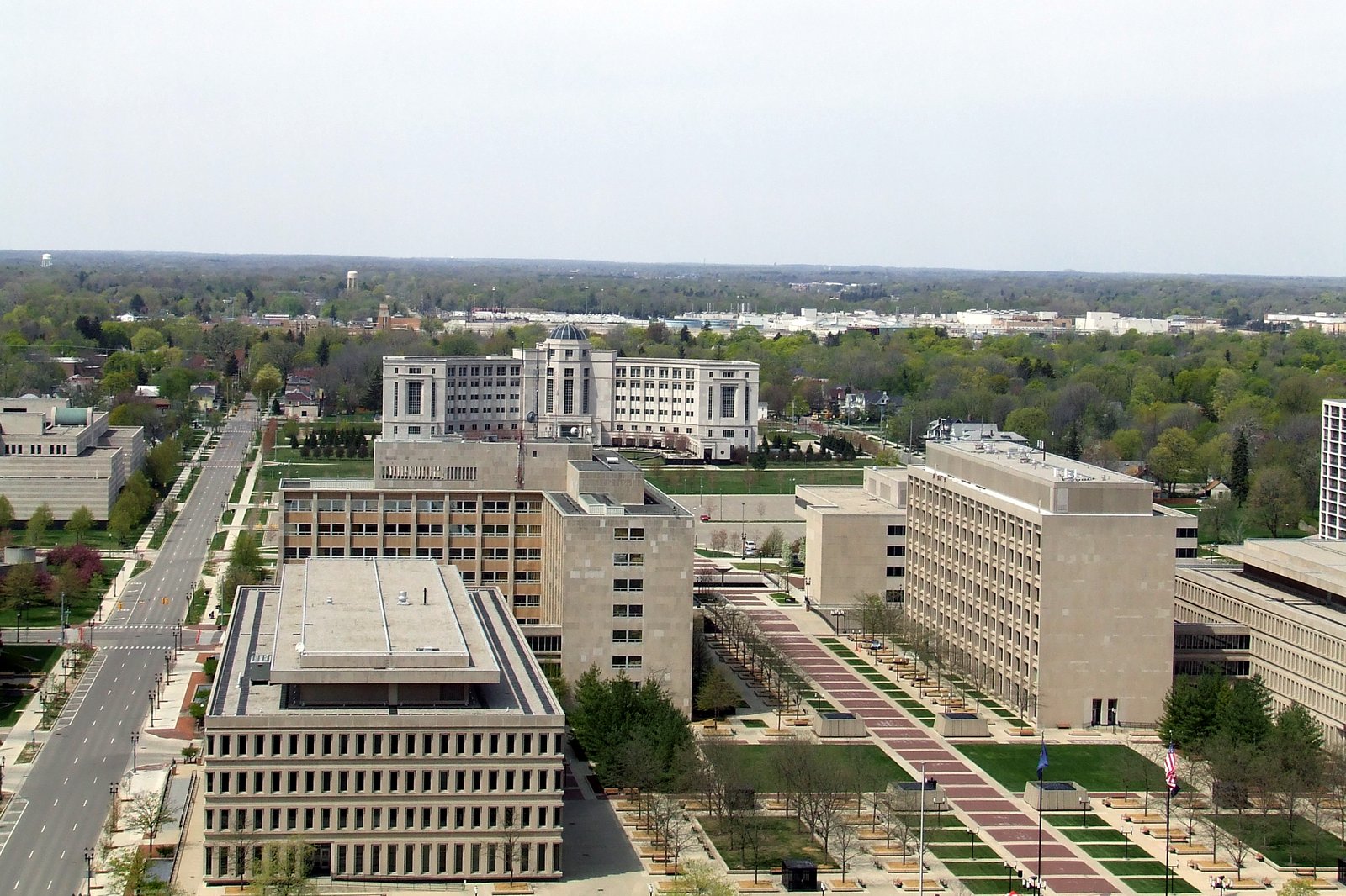 An aerial view of large pale stone state office buildings around a long brick-paved mall with trees, with a domed white building beyond.