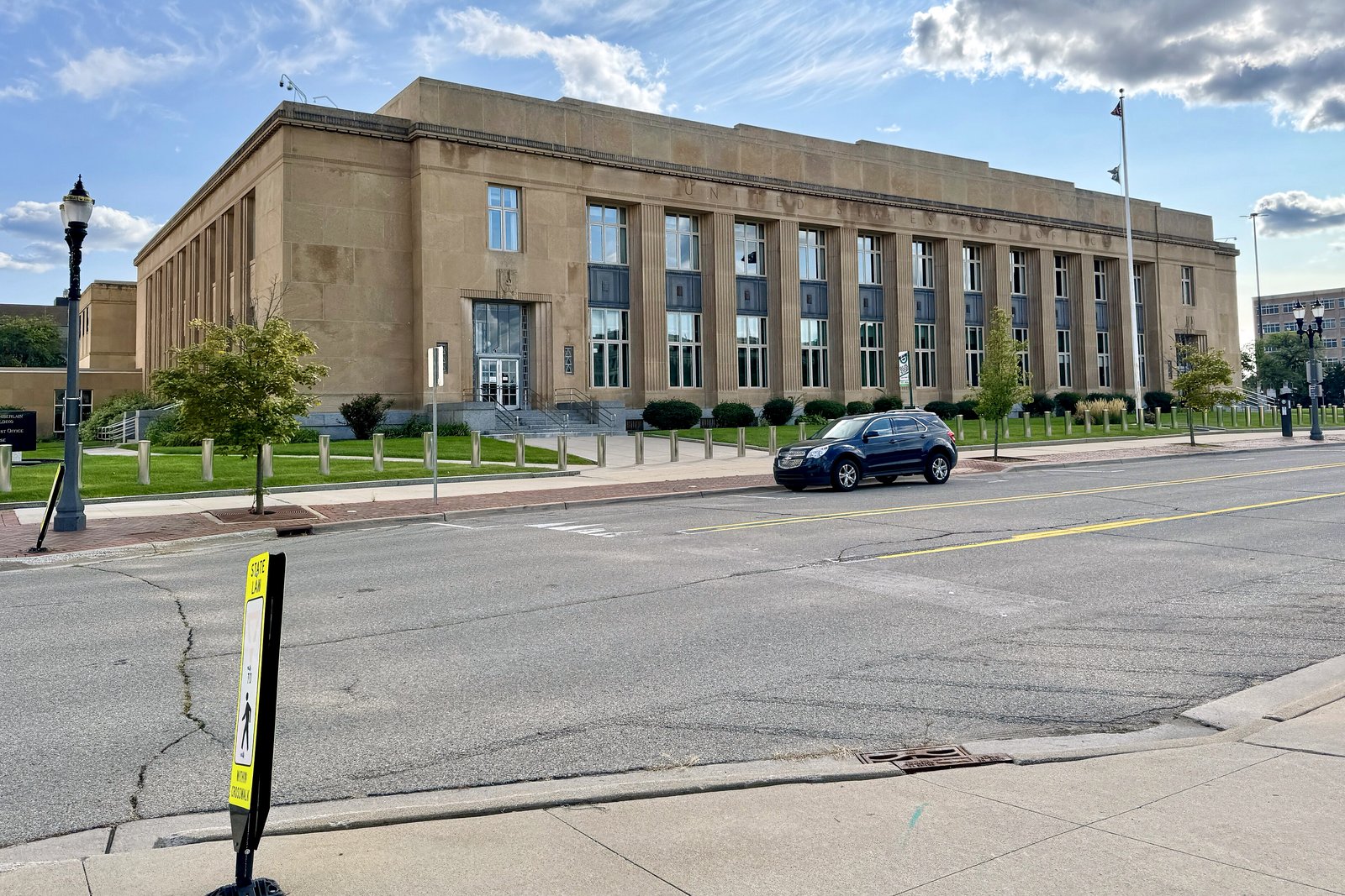 A pale stone federal building with tall vertical windows and carved panels stands on a downtown corner beside trees and a parked car.