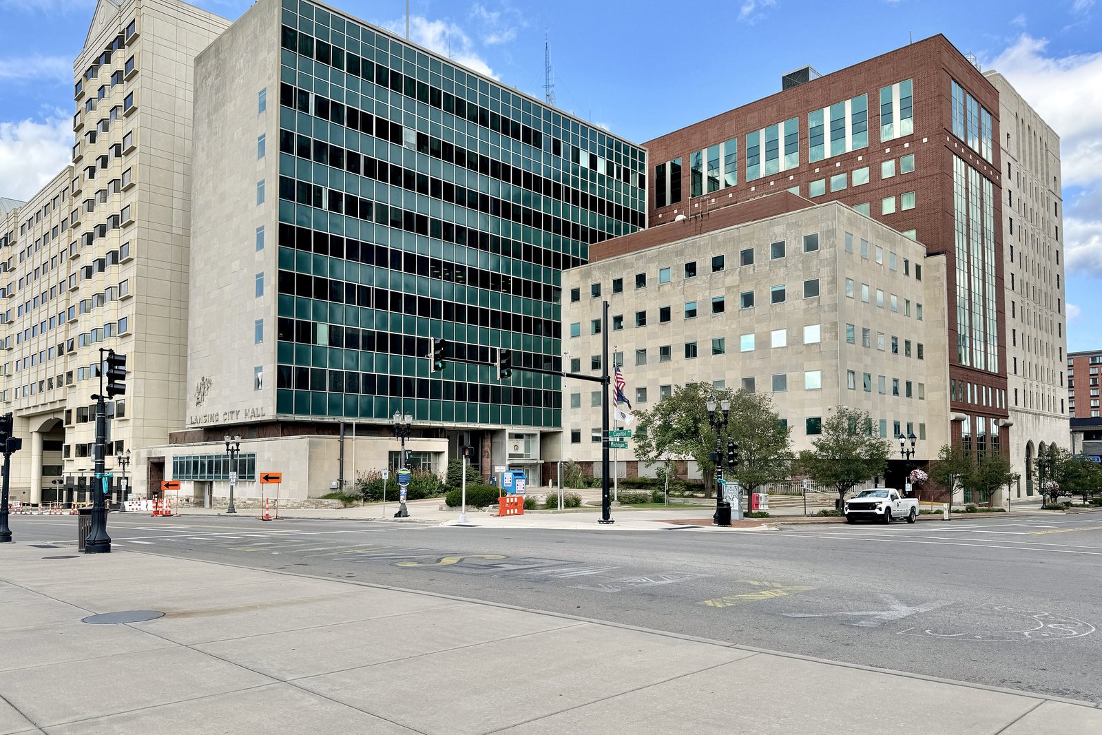 Lansing City Hall, a mid-century glass and stone office building, at a downtown intersection under a partly cloudy sky.