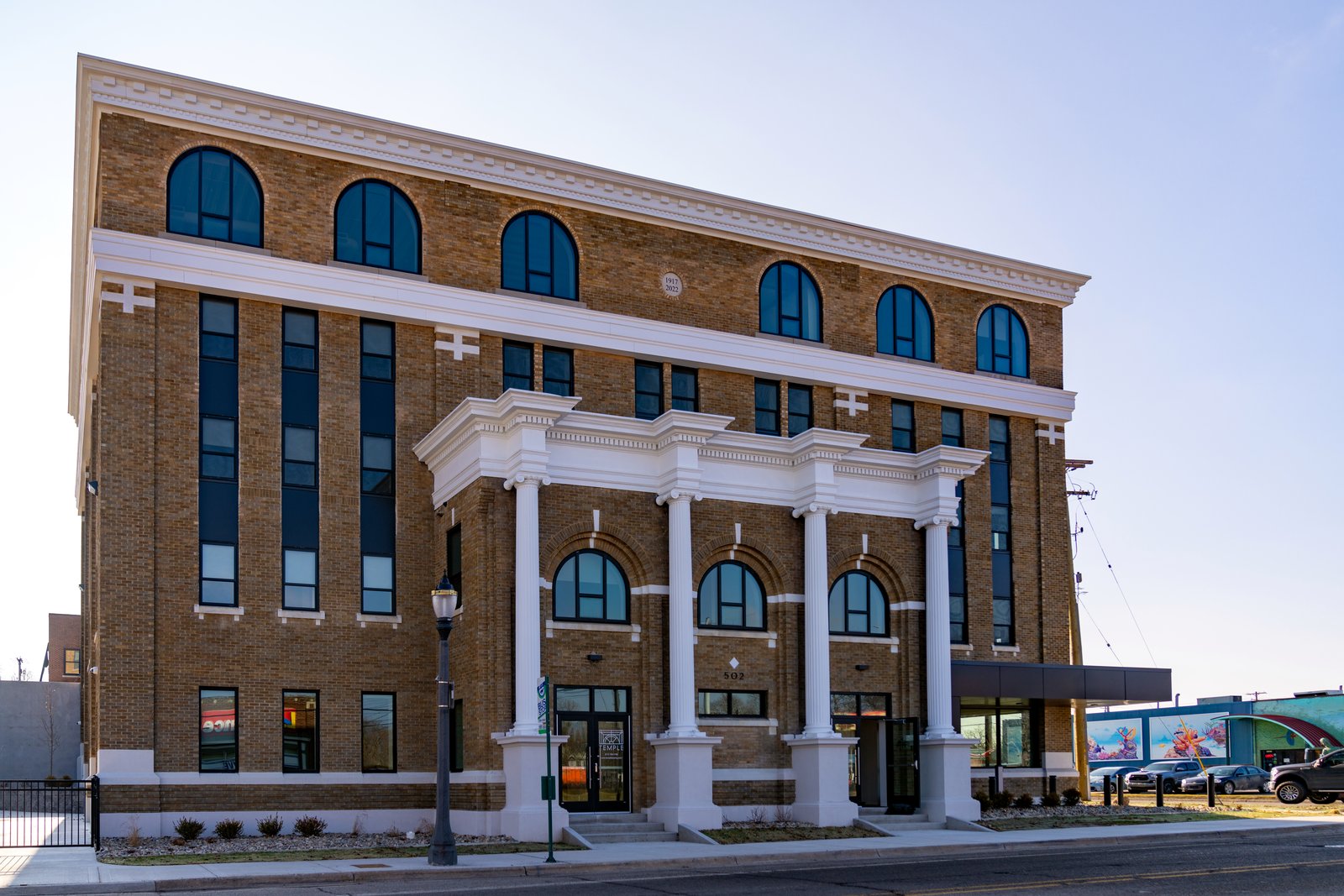 A four-story tan brick building with tall arched windows and a white columned entrance stands on a street corner under a pale sky.