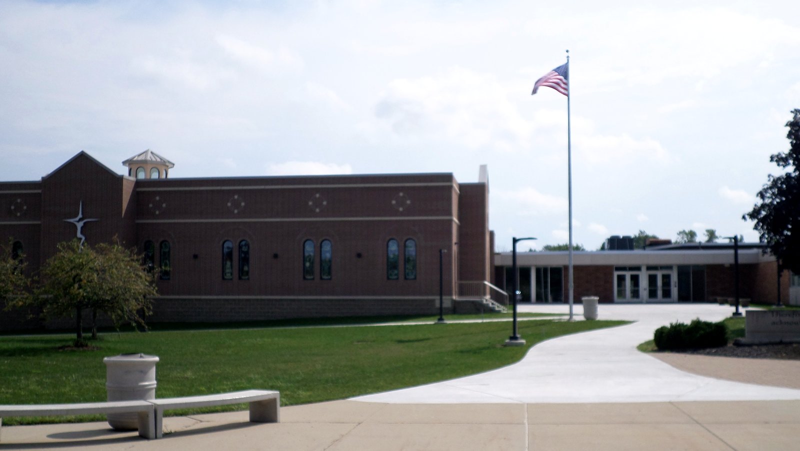 A long red-brick school building with tall arched windows and a small cupola, an American flag on a tall pole, a curving sidewalk and green lawn in front under a cloudy sky.