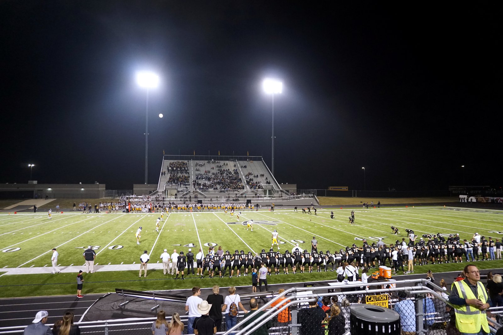 High school football teams line up for an opening kickoff on a lit field at night, with a band on the sideline and bleachers behind it.
