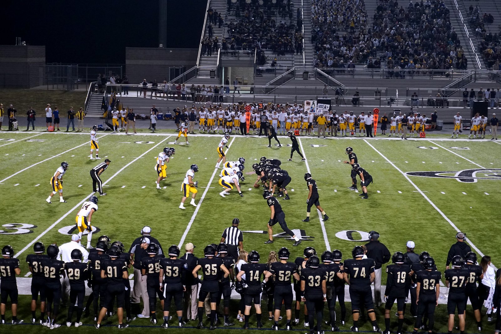 High school football players in gold and white line up against a team in black at night, with players in black jerseys along the near sideline and full bleachers behind.