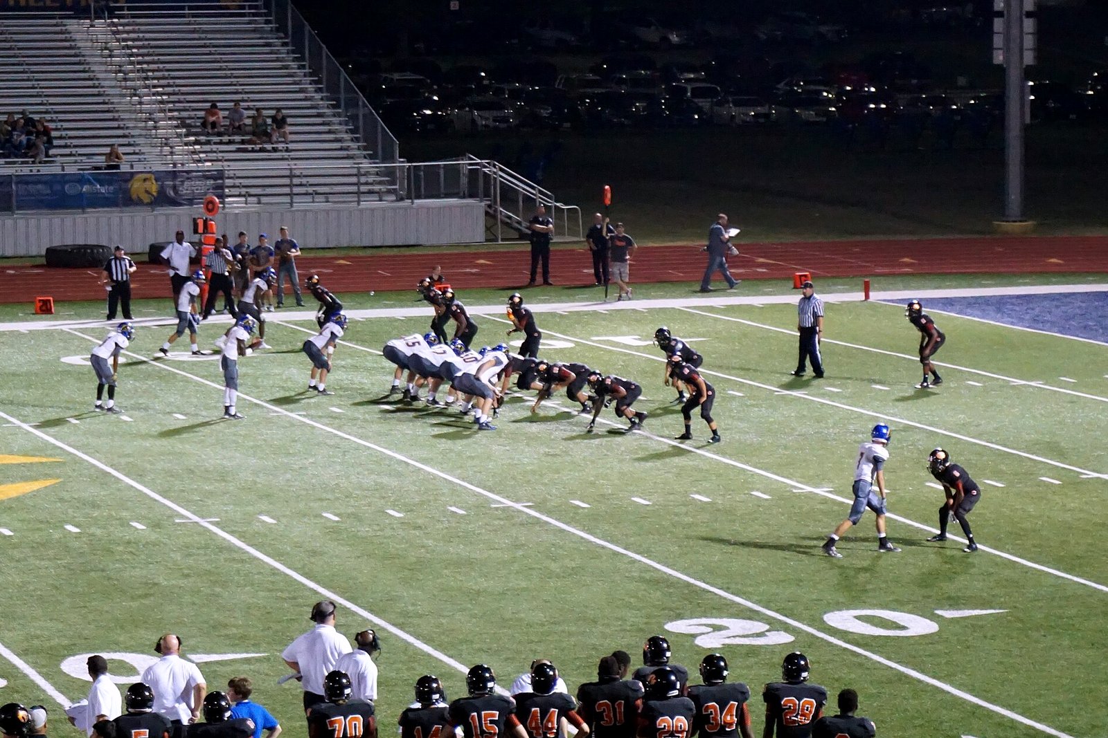 High school football teams in white and dark jerseys run a play on an artificial-turf field at night as players on the sideline watch.