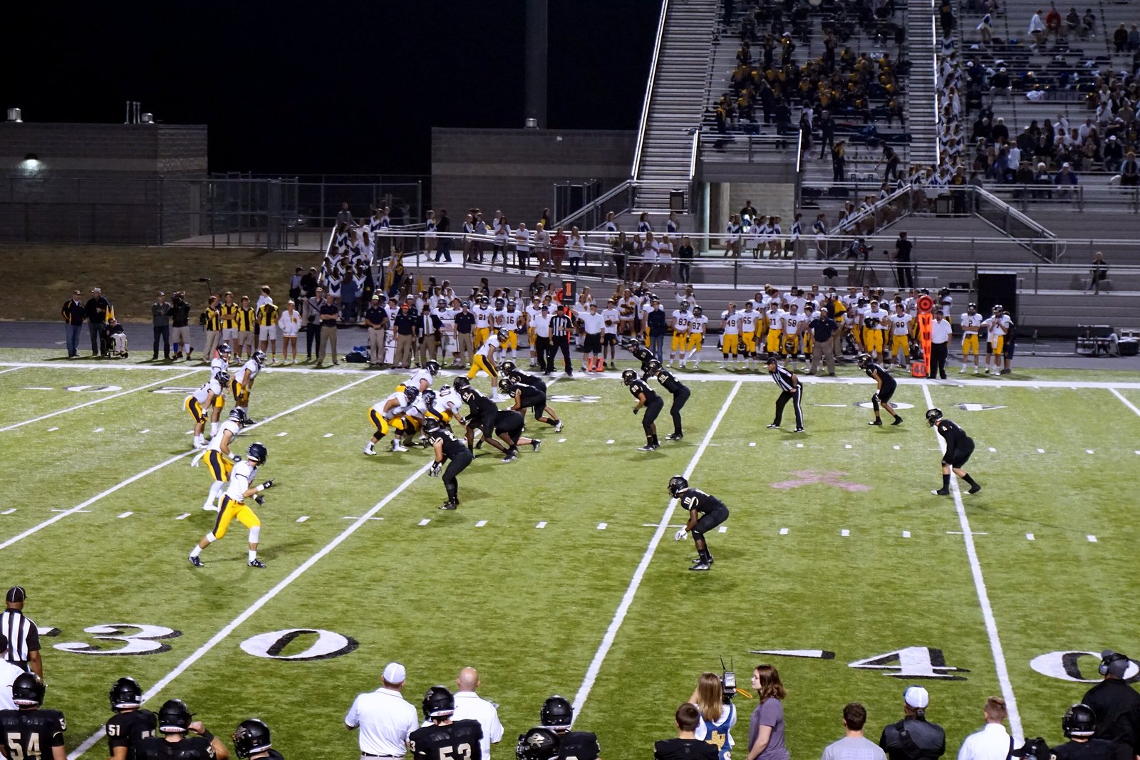 A high school football team in white and gold lines up on offense on a green turf field as players and coaches watch from the sideline in front of the grandstand.