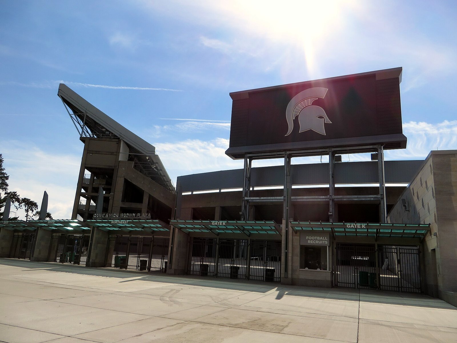 The south end of Spartan Stadium in East Lansing on a sunny day, with a large dark video board bearing the white Spartan helmet logo above gated entrances, a tall concrete upper-deck support on the left and an empty plaza in front.