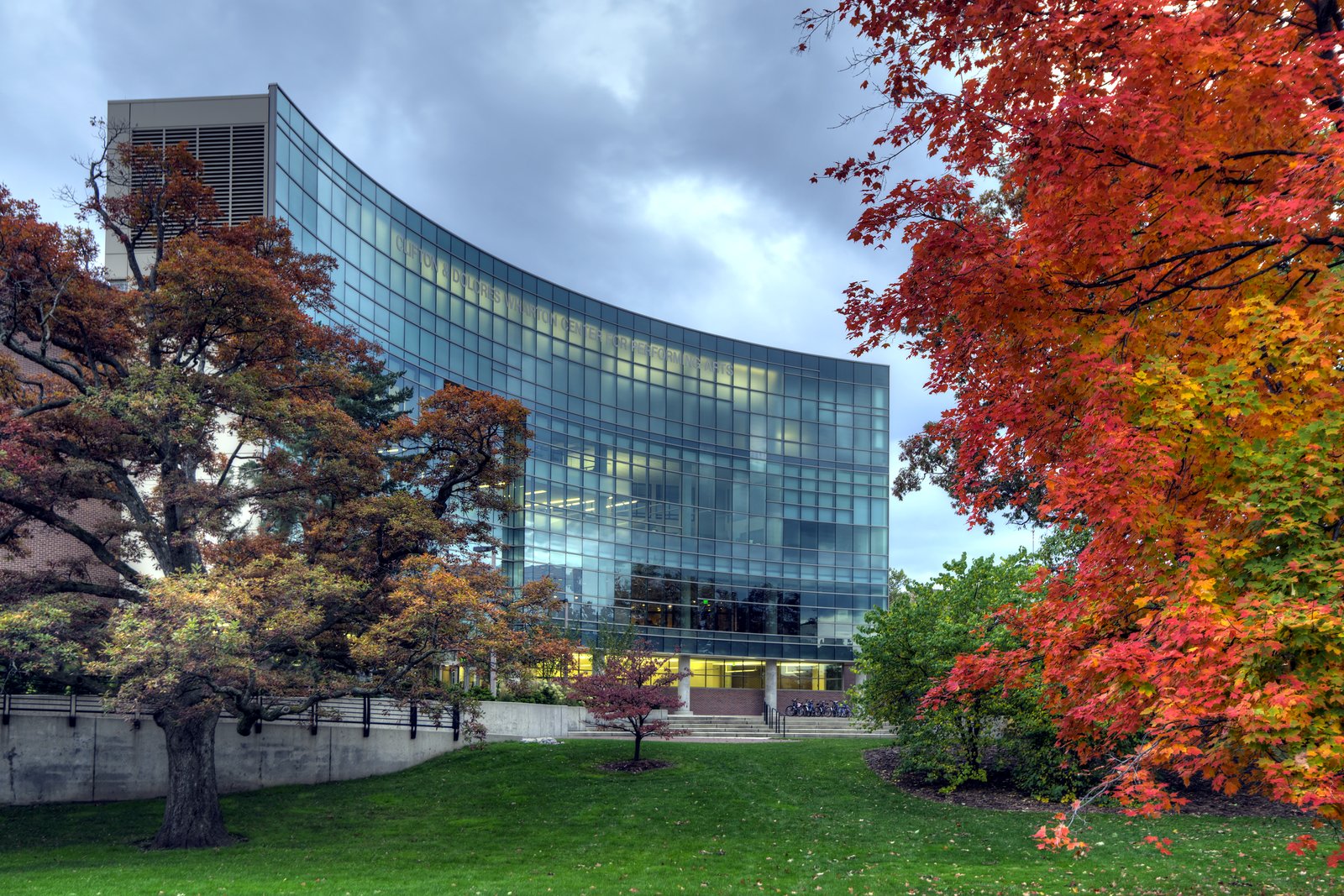 The curved glass front of the Wharton Center for Performing Arts at Michigan State University, framed by trees in red, orange and gold fall color over a green lawn.