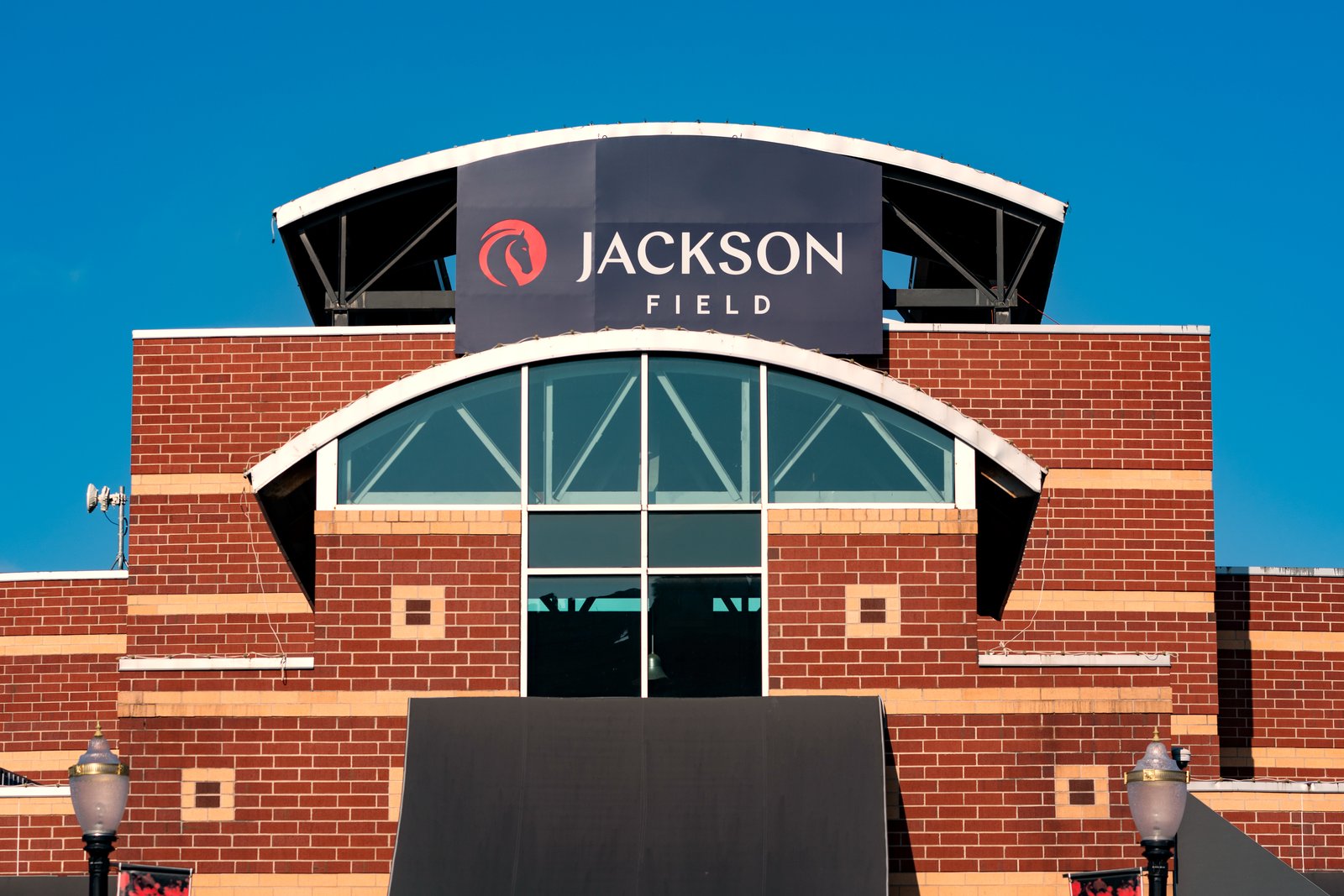 The brick entrance of Jackson Field in Lansing, with an arched window and a dark Jackson Field sign under a curved roof against a clear blue sky.