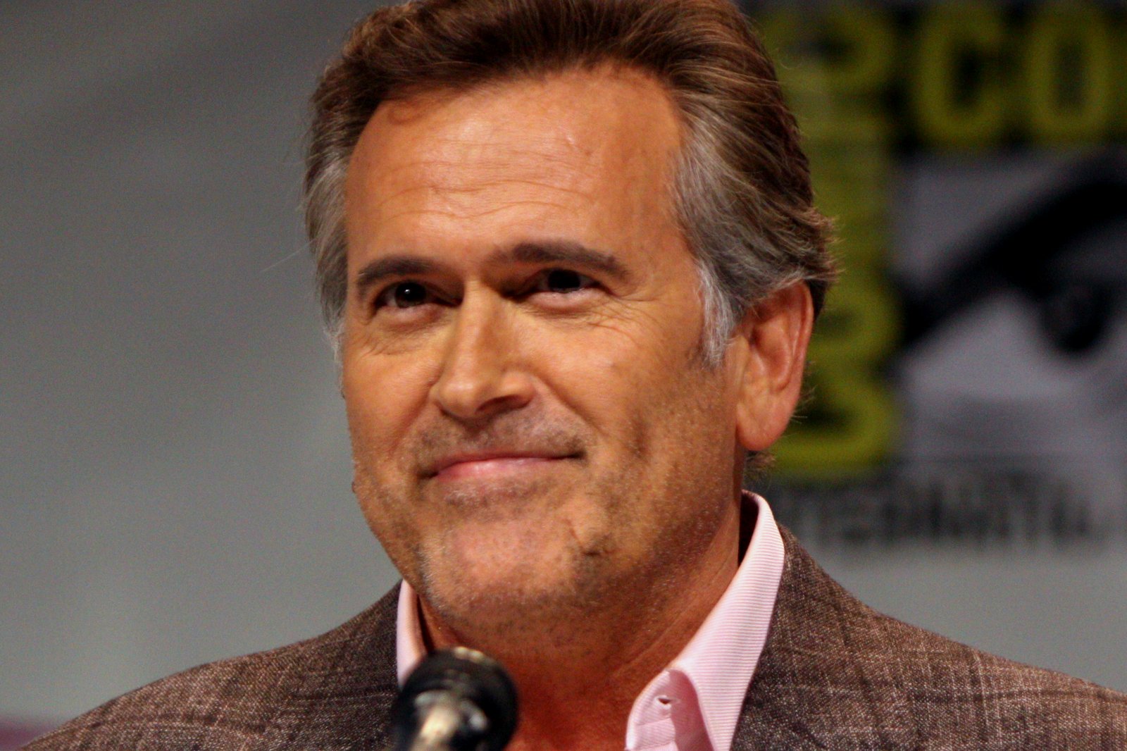 A smiling man with swept-back gray hair, in a brown checked blazer and pink shirt, speaks at a microphone during a convention panel.