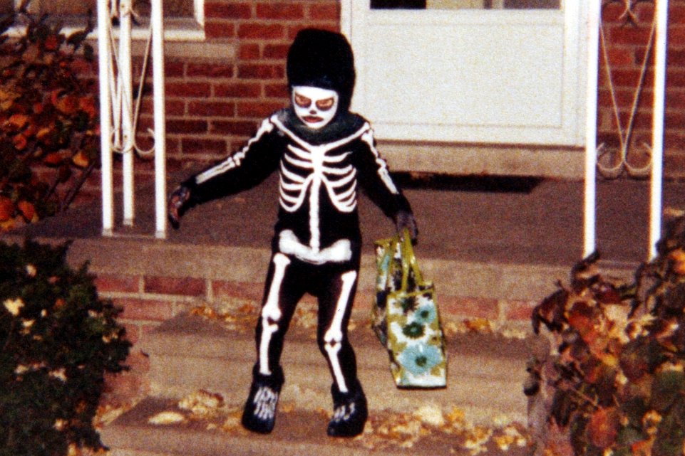 A child in a black skeleton costume and mask standing on brick front steps decorated with fall leaves and a jack-o-lantern bucket.