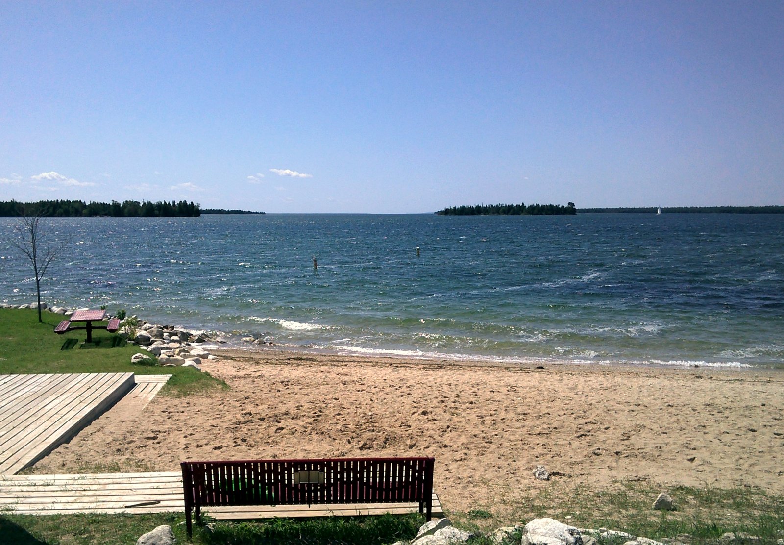 A sandy beach with a wooden bench and boardwalk on Hessel Bay, with blue water and low wooded islands on the horizon under a clear sky.