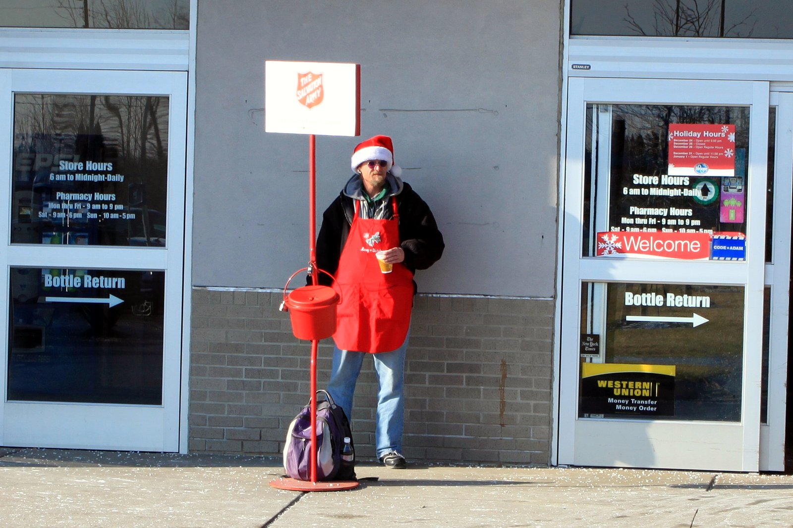 A bell ringer in a red apron stands beside a Salvation Army red kettle at a supermarket entrance.