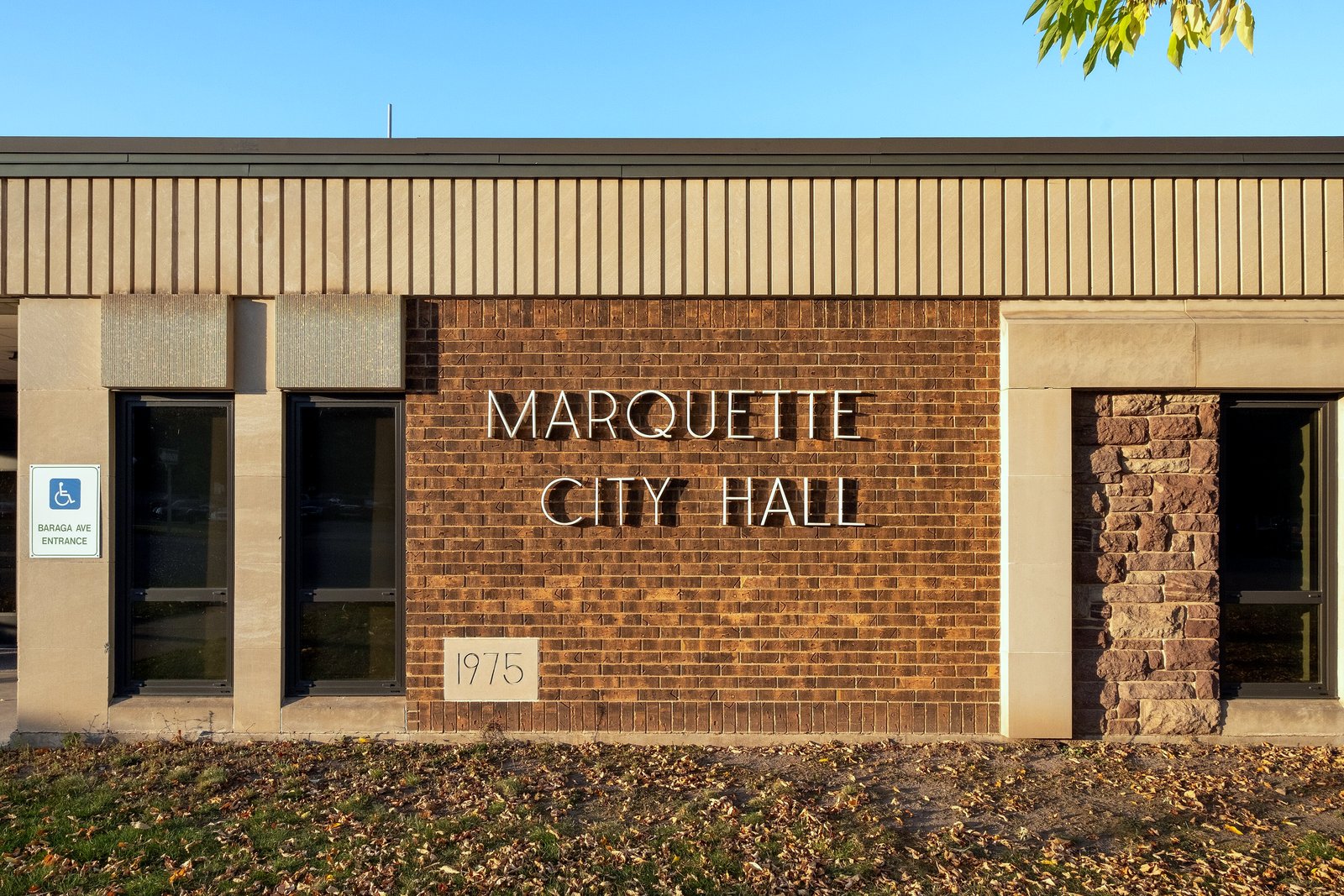 A tan brick wall with raised letters reading Marquette City Hall and a small 1975 datestone, beside windows and a stone pillar, with fallen leaves on the grass in front.