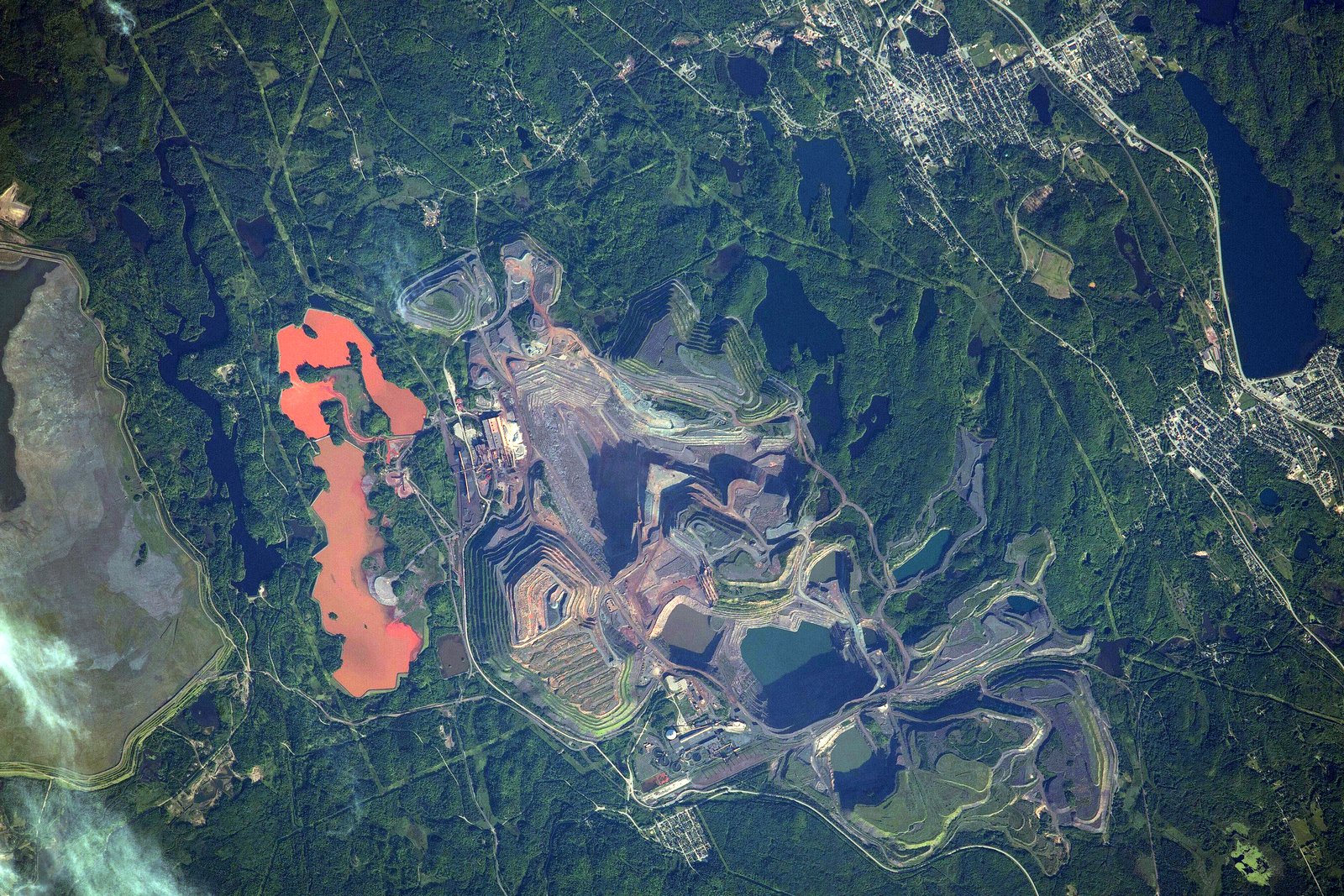 A view from space of reddish open-pit mine areas and gray tailings among green forest and lakes.