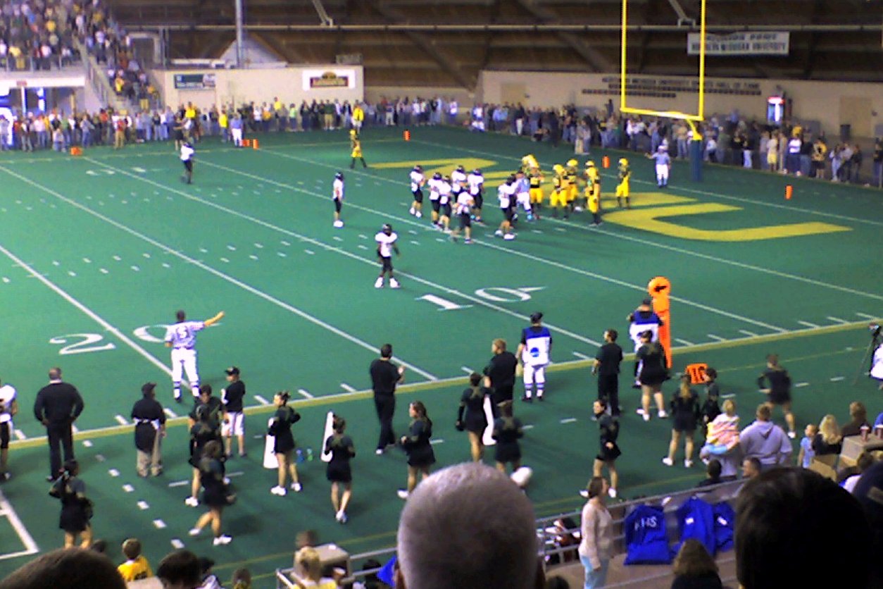 A college football game on green artificial turf inside a domed stadium with a wooden roof, with players on the field and fans lining the sideline.