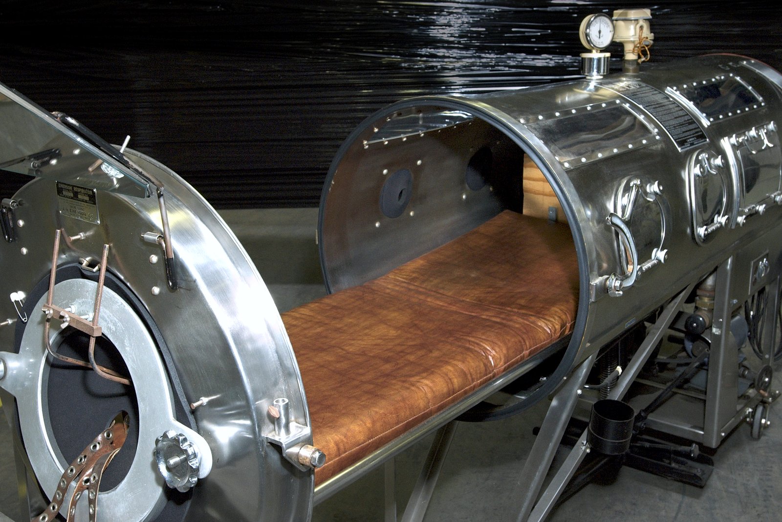 An open, cylindrical metal respirator known as an iron lung, with a padded bed slid partly out of the chamber.