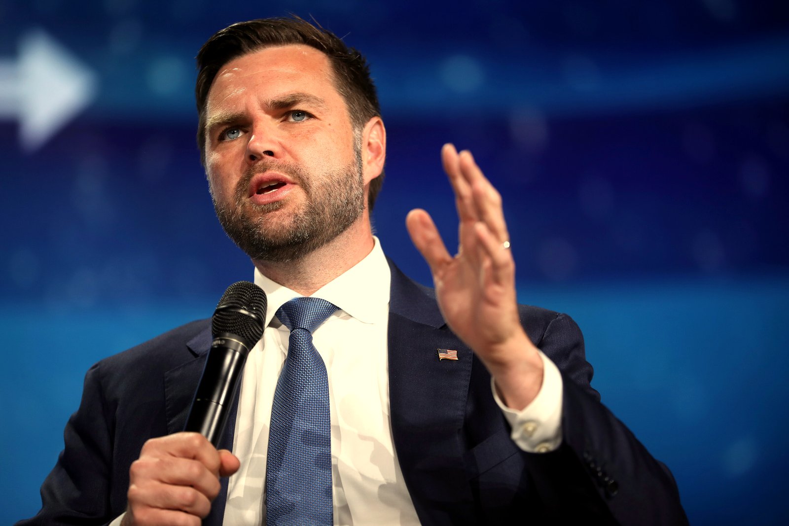 JD Vance, in a navy suit, white shirt and blue tie, speaks into a handheld microphone and raises his right hand while seated in front of a blue stage backdrop.