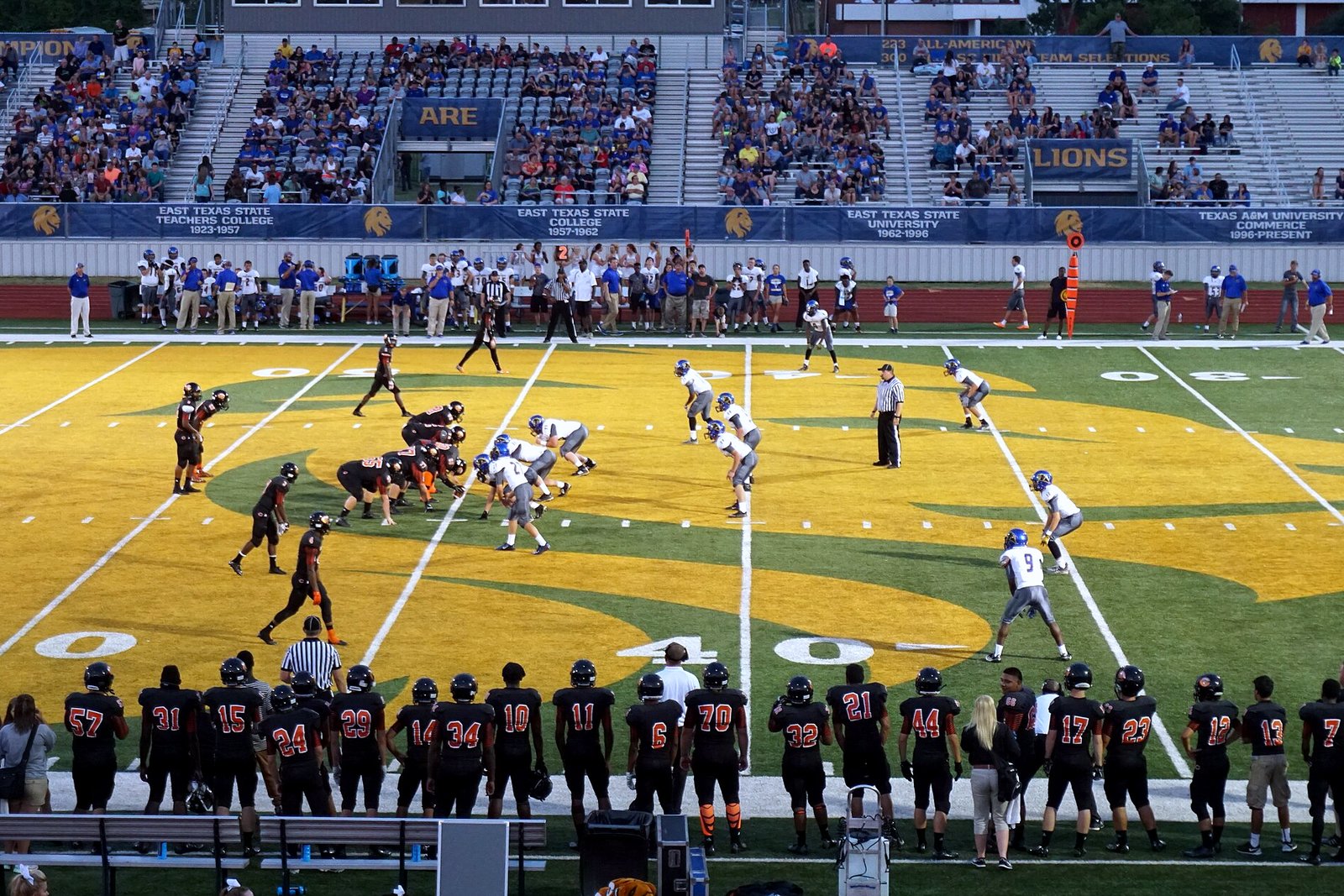 High school football players in white and dark jerseys line up at the line of scrimmage on a gold-and-green field in front of crowded grandstands.