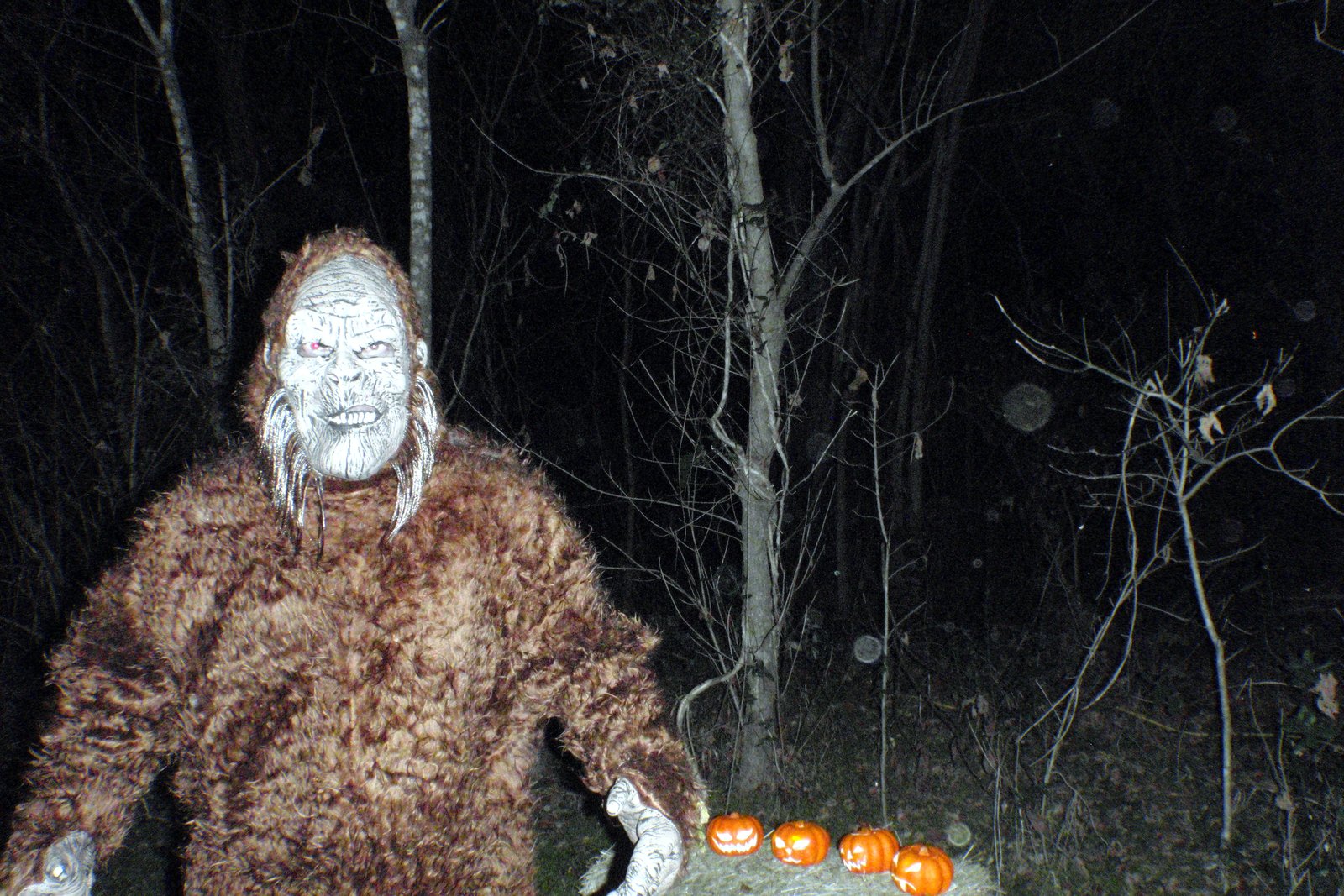 People in Halloween costumes ride a hay wagon at night past lit buildings during a haunted hayride.