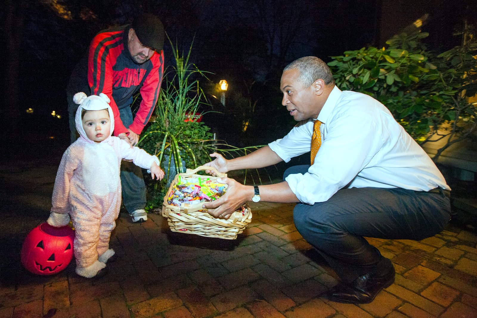 A small child in a white bunny costume holds a pumpkin bucket at night as a smiling man in a white shirt and tie crouches to offer candy from a basket.