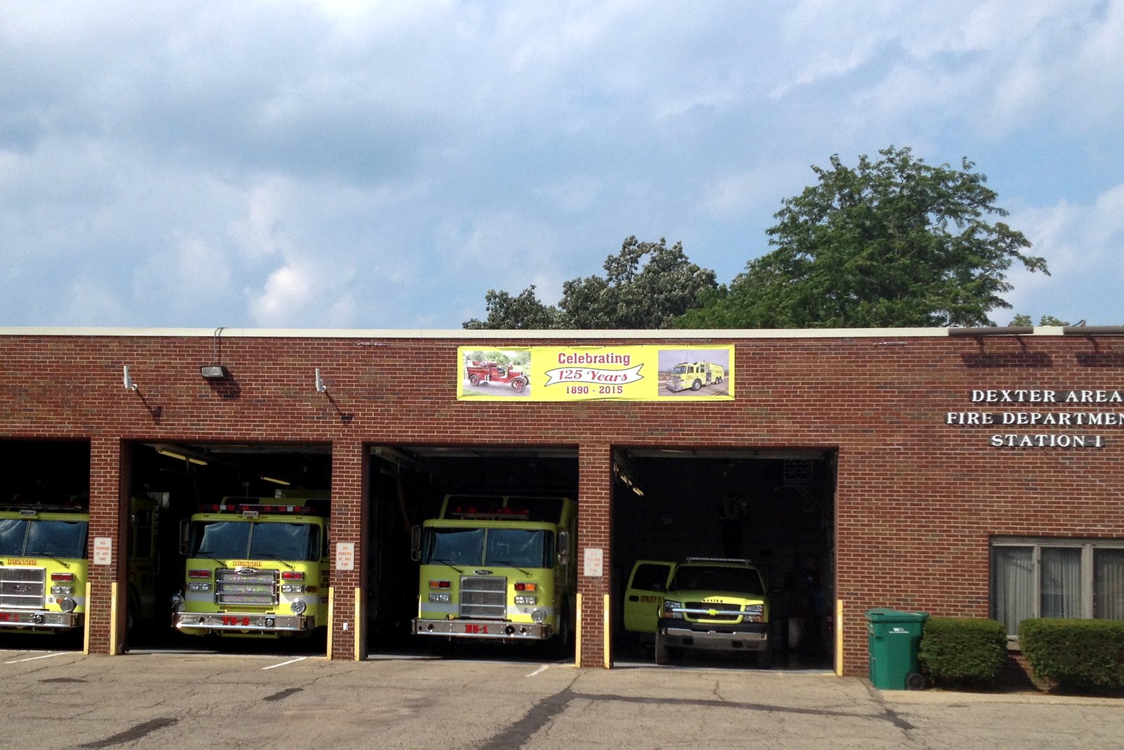 Four yellow-green fire trucks sit in the open bays of a one-story brick fire station under a partly cloudy sky.