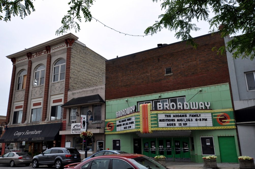 A downtown street of two- and three-story brick storefronts with a green-and-red theater marquee reading Broadway, cars parked at the curb and tree branches hanging over the sidewalk.