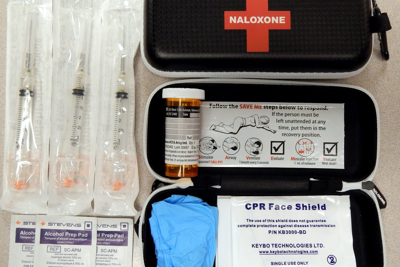 An open black naloxone kit holds syringes, vials, gloves, alcohol swabs and a CPR face shield.