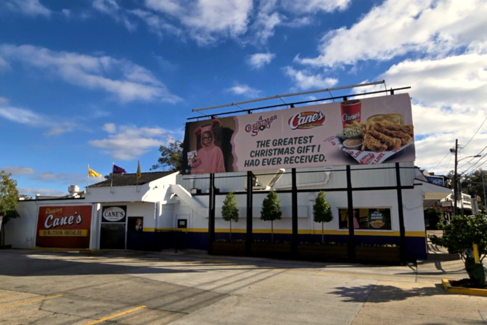 A white Raising Cane’s restaurant building with a red sign and a large billboard on the roof, beside a drive-through lane under a blue sky with clouds.